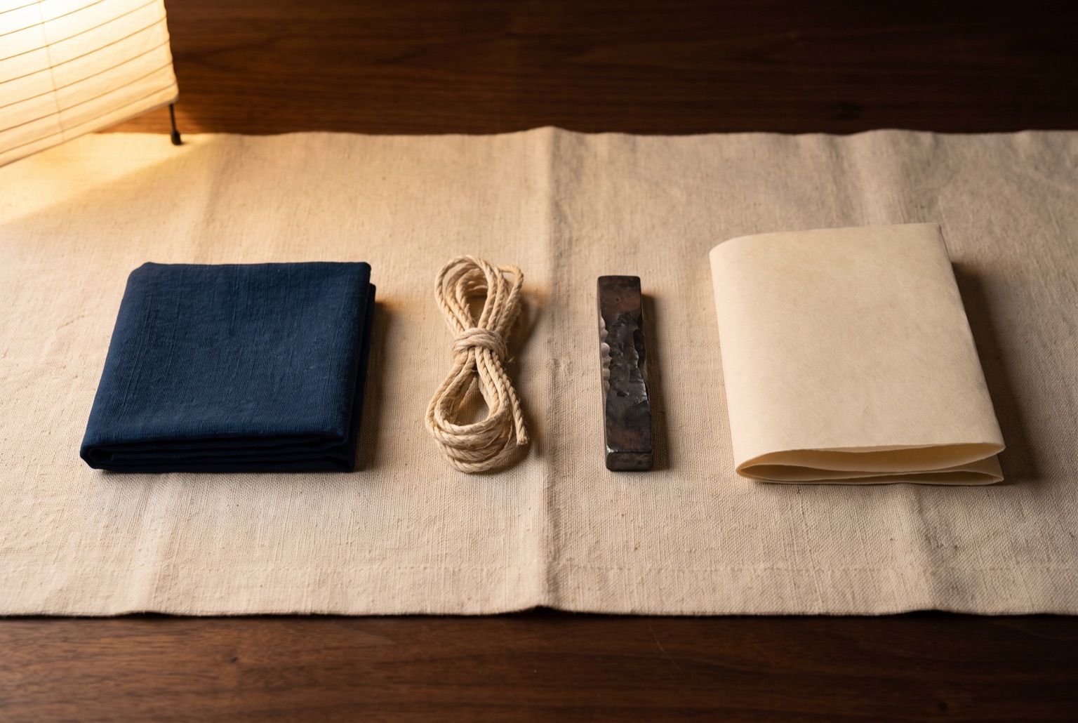 Four material samples laid in a row on pale hemp cloth: folded indigo cotton, a coil of hemp rope, a bar of forged iron, and a sheet of unbleached washi.