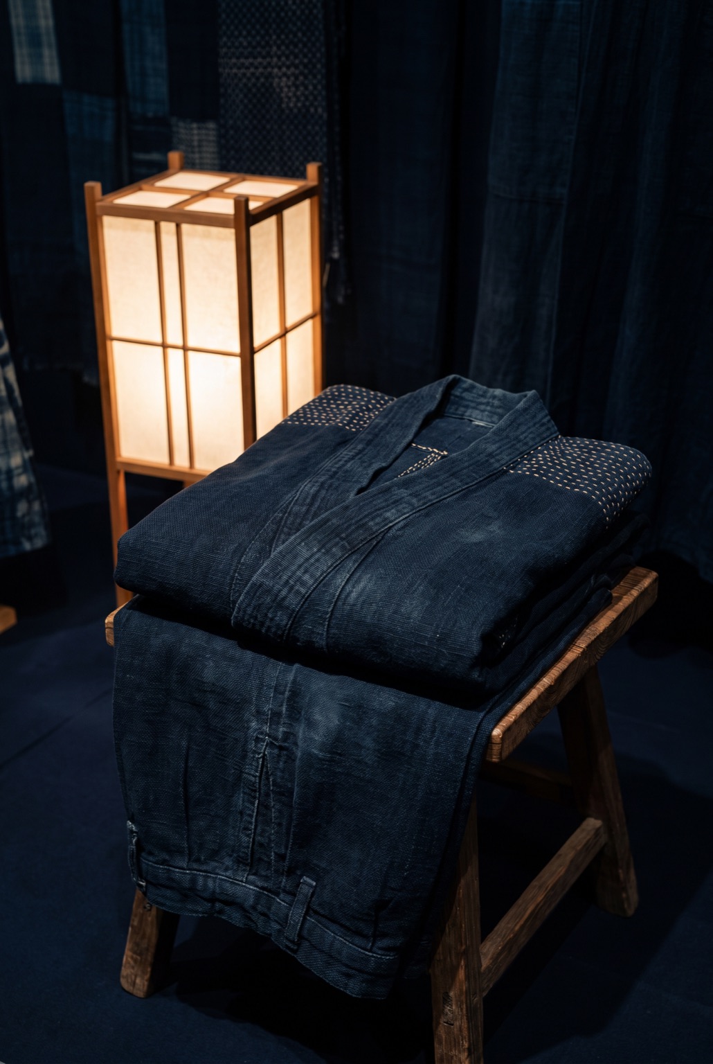 An indigo-dyed cotton work jacket and trousers with sashiko stitching, folded into a neat rectangle on a plain wooden stand.